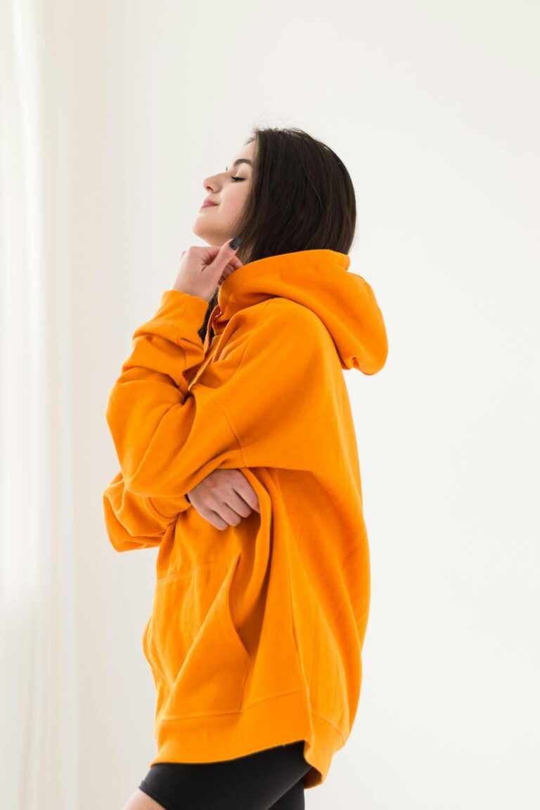 Young woman in an orange oversized hoodie posing in a bright studio setting.